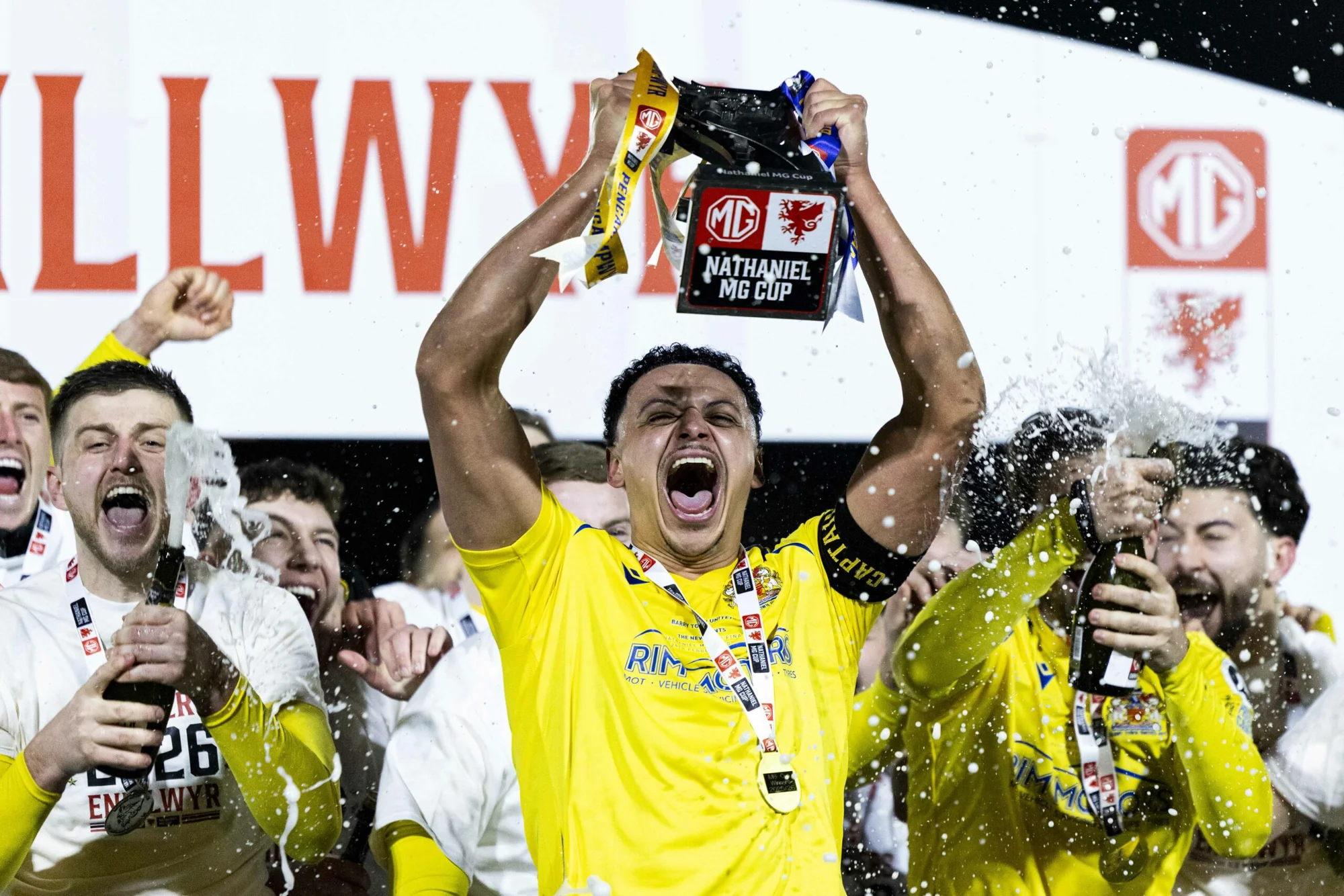 Barry Town celebrating their 2026 Nathaniel MG Cup win, with the trophy raised above yellow-shirted players